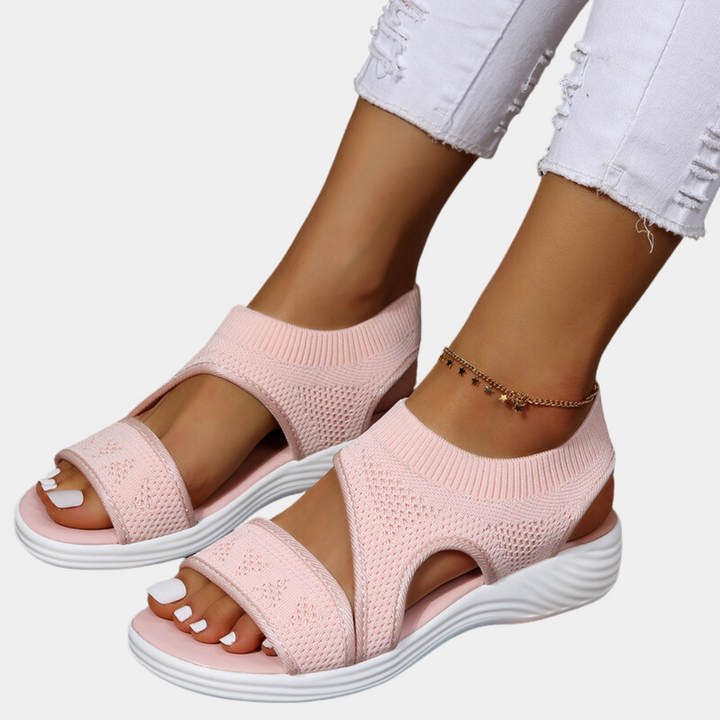 Roze gebreide sandalen met dikke witte zool, gedragen door vrouw met witte jeans en enkelbandje. Comfortabele zomerschoenen voor dames.