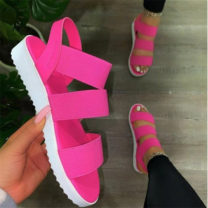 Felroze sandalen met witte zool, elastische banden en open teen, gedragen door een persoon op een houten vloer. Trendy zomerschoenen voor dames.