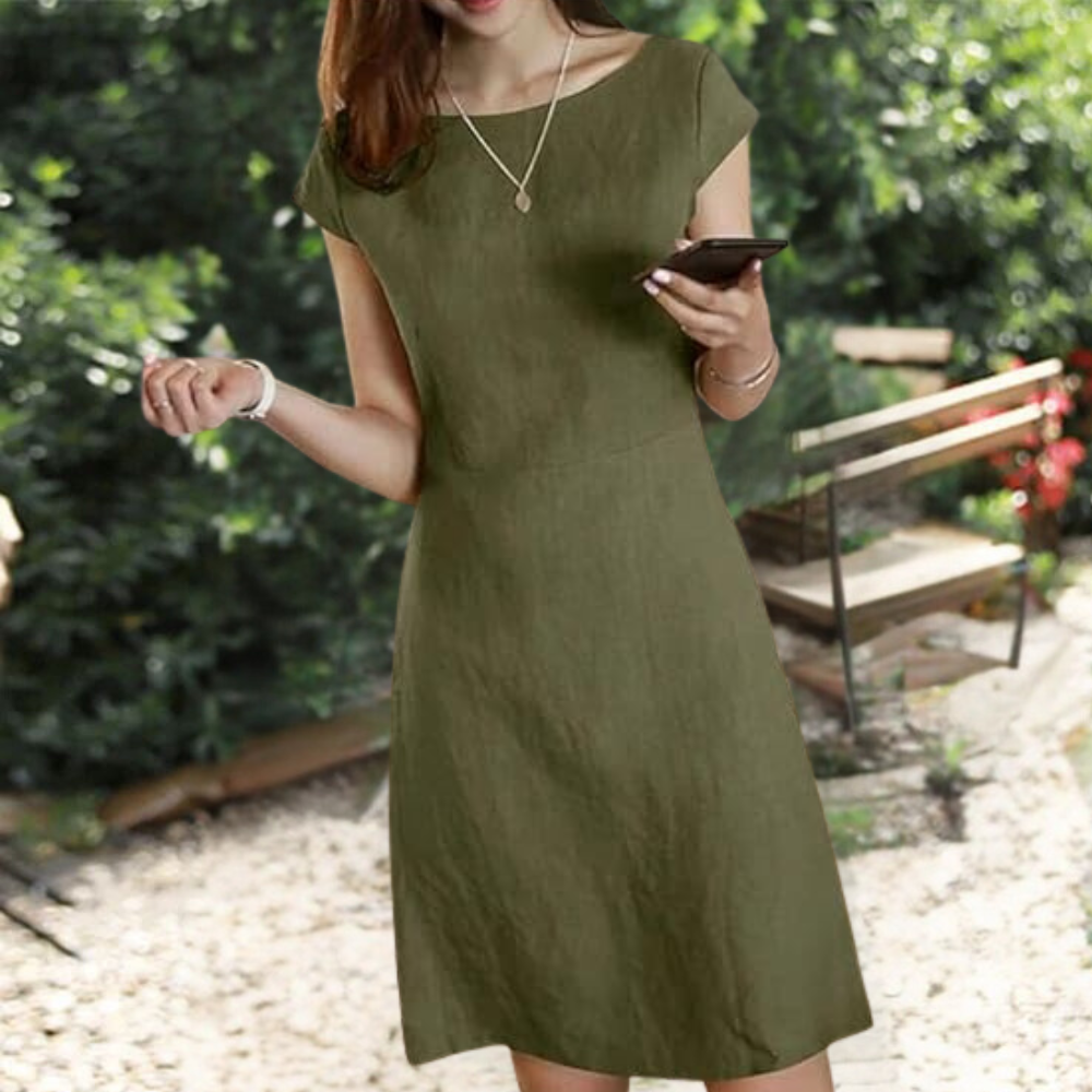Vrouw in groene Henka-jurk, zomerkleding, buiten in tuin, smartphone in hand, casual mode, comfortabele damesjurk, trendy outfit.