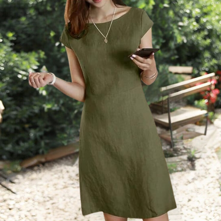 Vrouw in groene Henka-jurk, zomerkleding, buiten in tuin, smartphone in hand, casual mode, comfortabele damesjurk, trendy outfit.