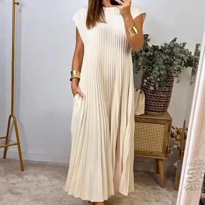 Vrouw in elegante, witte, geplooide maxi-jurk met mouwloze snit, gouden armbanden, naast houten kast met groene plant. Perfect voor zomermode.
