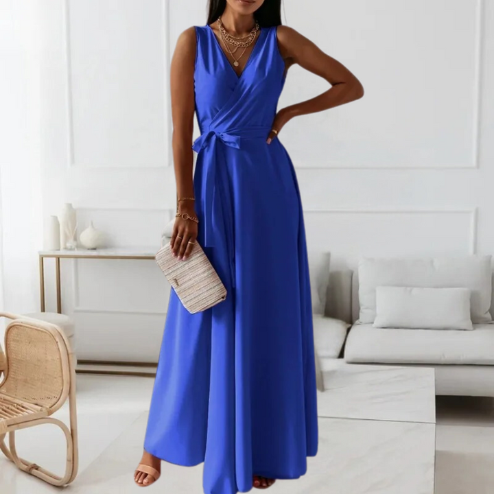 Vrouw in elegante blauwe maxi-jurk met V-hals en strik, staand in moderne woonkamer. Accessoires: beige clutch en gouden sieraden.