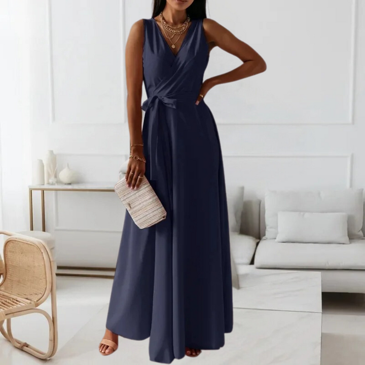 Elegante marineblauwe maxi-jurk met V-hals en strikceintuur, perfect voor avondfeesten. Draagster houdt een beige clutch vast in een moderne woonkamer.