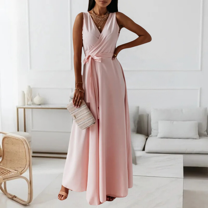 Elegante roze maxi-jurk met V-hals en strikceintuur, gedragen door een vrouw. Perfect voor zomerfeesten. Accessoires: beige clutch, gouden sieraden.