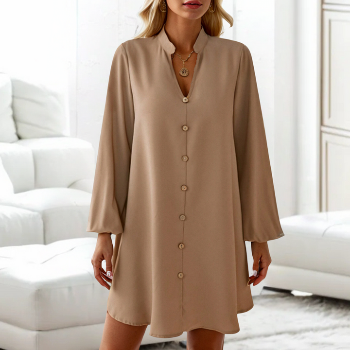 Vrouw in beige overhemdjurk met knopen, lange mouwen, staande in moderne woonkamer. Elegante damesmode, casual chique stijl.