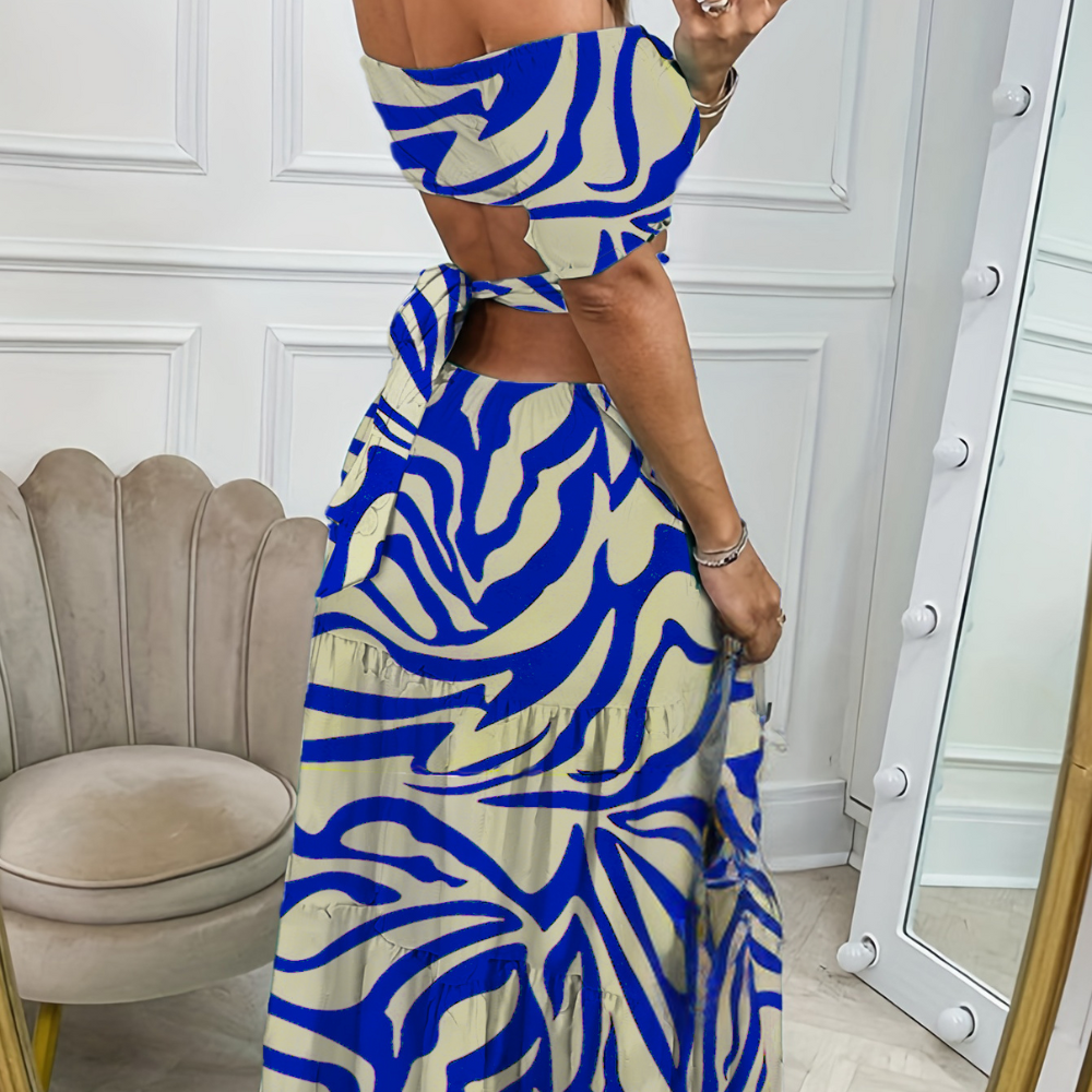 Vrouw in elegante blauwe en beige jurk met abstract patroon, off-shoulder en open rug, poseert voor spiegel in stijlvolle kamer.