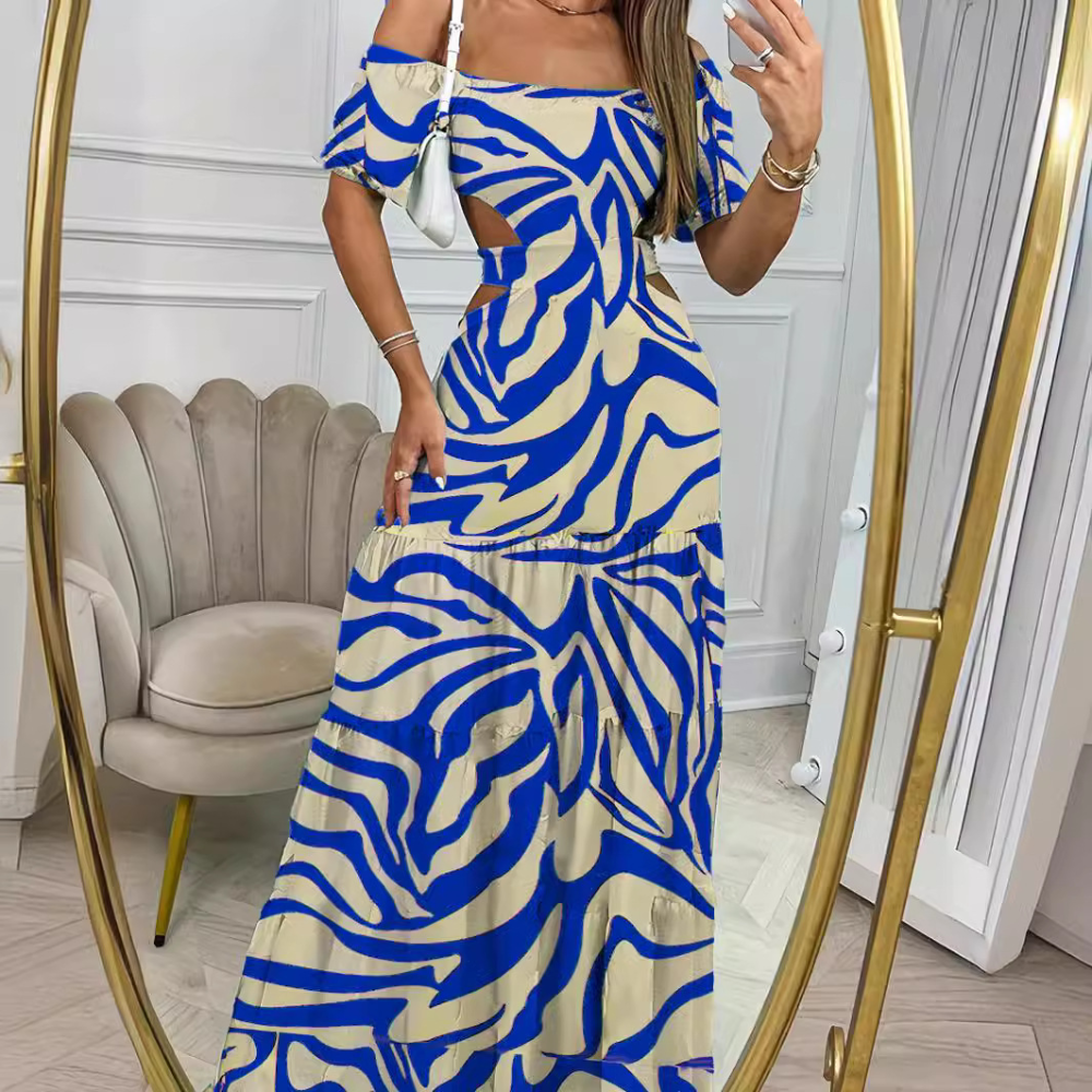 Vrouw in blauwe en beige maxi-jurk met abstract patroon, poserend voor een spiegel in een elegante kamer. Trendy zomerjurk, off-shoulder stijl.