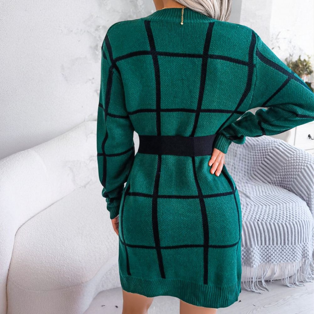 Vrouw in groene geruite trui-jurk met zwarte riem, staand in moderne woonkamer. Stijlvolle wintermode, comfortabele en trendy outfit.