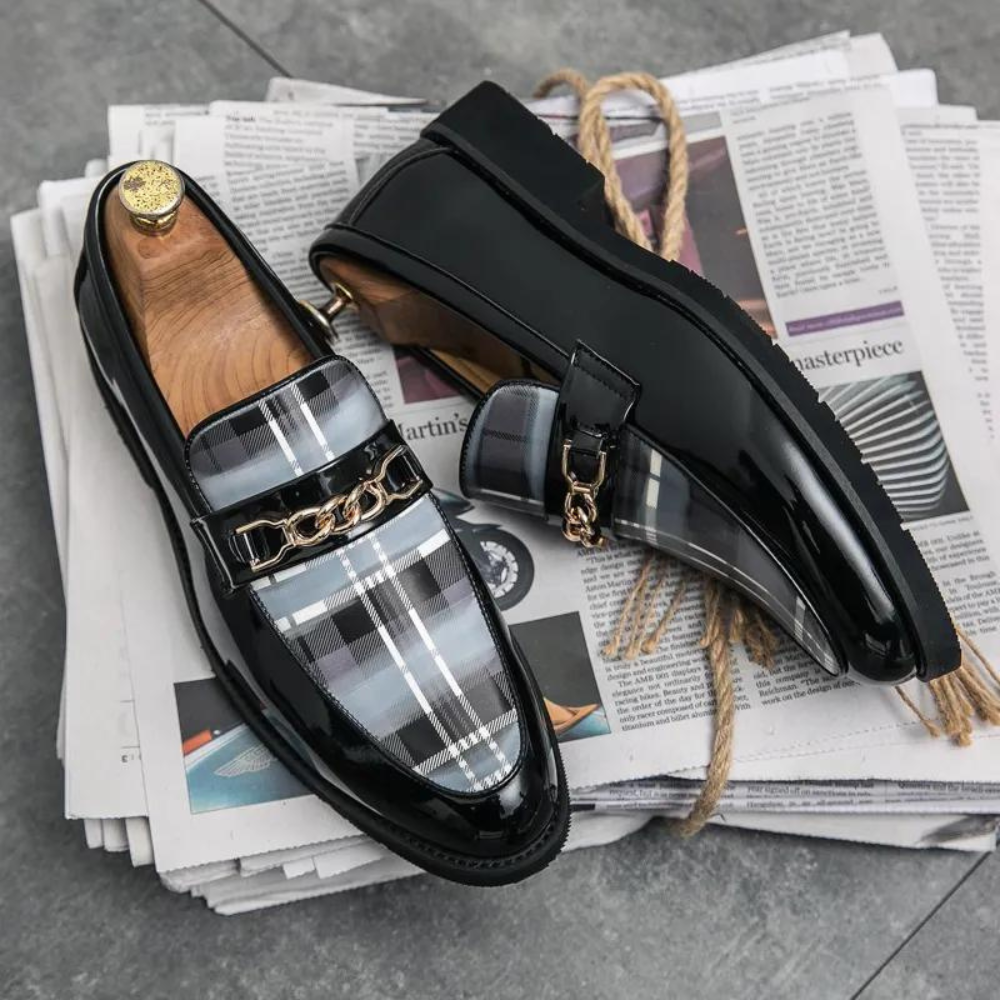 Zwart gelakte leren loafers met geruit patroon en gouden kettingdetail, stijlvolle herenschoenen op kranten, luxe modeaccessoire.
