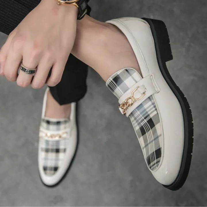 Witte leren loafers met zwart-wit geruit patroon en gouden kettingdetail, gedragen door een persoon op een grijze achtergrond. Stijlvolle herenschoenen.