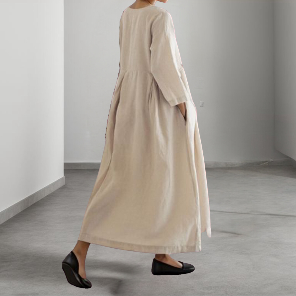 Vrouw in lange, beige linnen jurk met driekwart mouwen, staand in minimalistische ruimte. Comfortabele, stijlvolle mode voor dames.