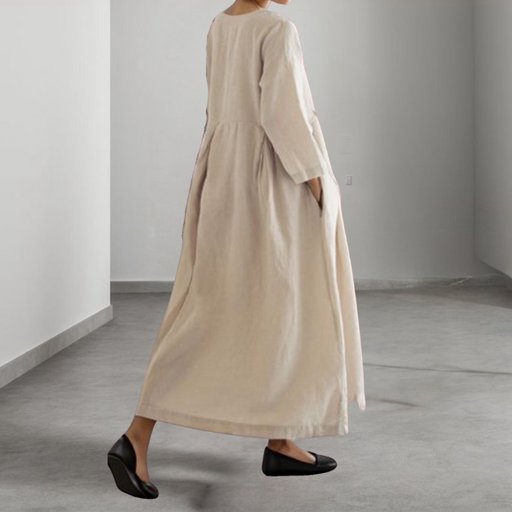 Vrouw in lange, beige linnen jurk met driekwart mouwen, staand in minimalistische ruimte. Comfortabele, stijlvolle mode voor dames.