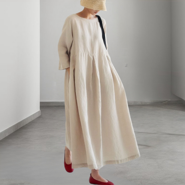 Vrouw in een lange, beige linnen jurk met driekwart mouwen, draagt een strohoed en rode schoenen, minimalistische achtergrond.