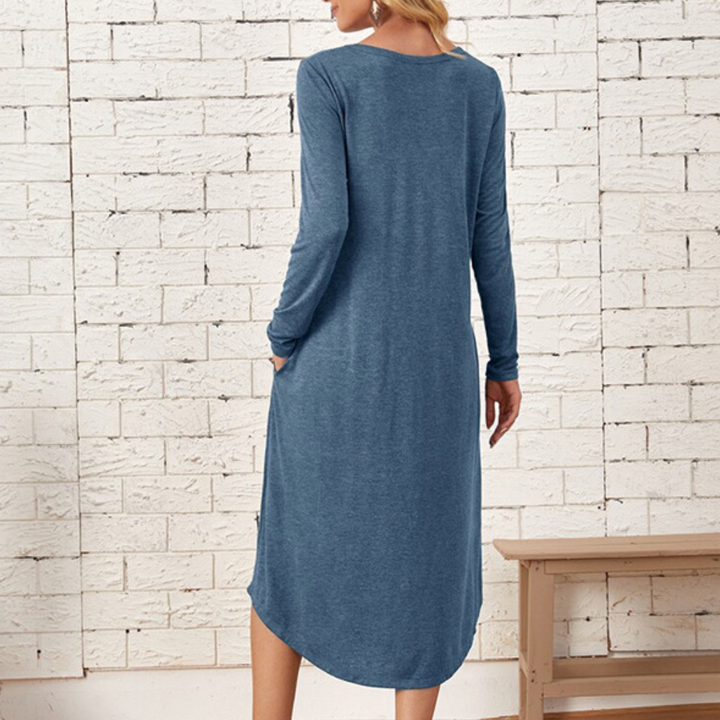 Vrouw in blauwe lange jurk met lange mouwen, staand voor witte bakstenen muur. Casual damesmode, comfortabele kledingstijl.