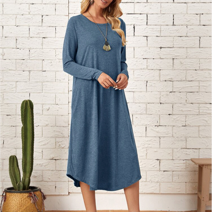 Blauwe lange jurk met lange mouwen, gedragen door een vrouw, voor een witte bakstenen muur. Casual damesmode, comfortabele pasvorm, herfstkleding.