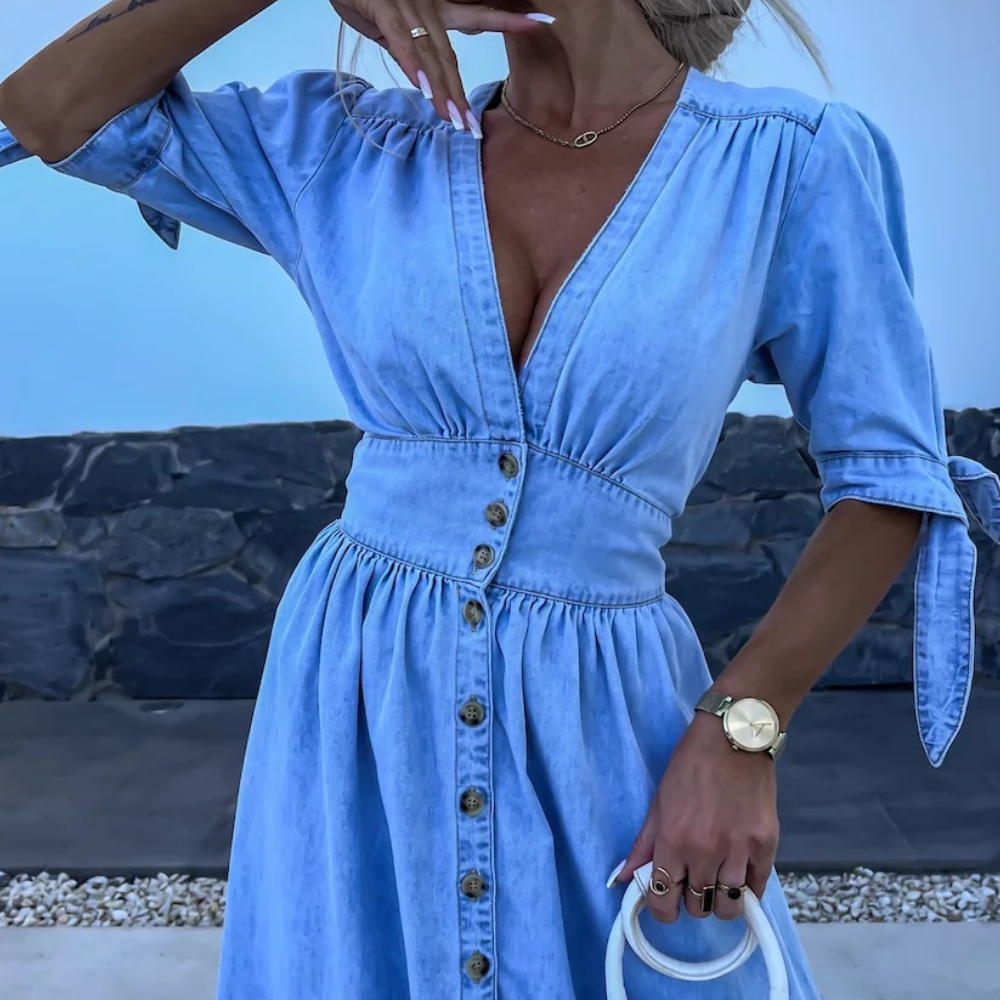 Vrouw in lichtblauwe denim jurk met knopen, korte mouwen en V-hals, draagt gouden horloge en witte handtas, tegen stenen achtergrond.