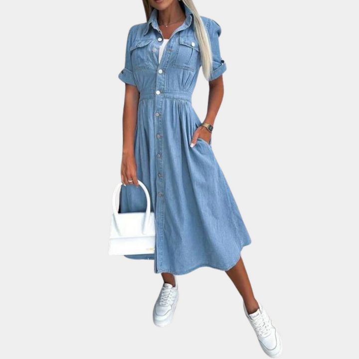 Vrouw in blauwe denim jurk met knoopsluiting, korte mouwen en zakken, gecombineerd met witte sneakers en een witte handtas. Modieuze casual outfit.