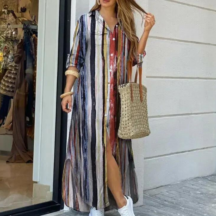 Vrouw in kleurrijke gestreepte maxi-jurk met lange mouwen, draagt een gevlochten tas en witte sneakers, poseert voor een winkel.