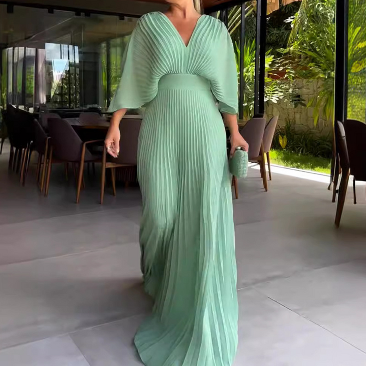 Vrouw in elegante groene plisséjurk met vleermuismouwen, wandelend in moderne eetkamer. Stijlvolle avondjurk, mode, luxe kleding.