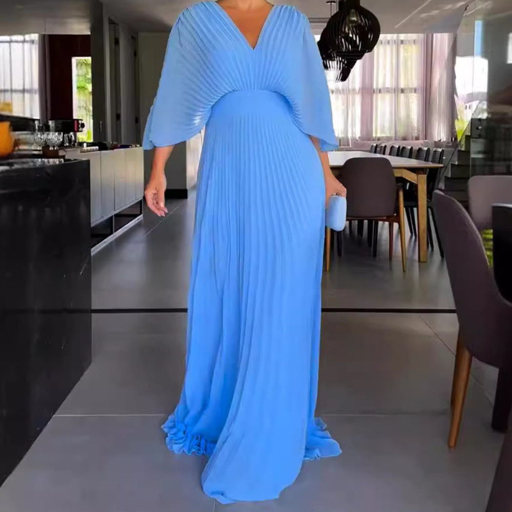 Vrouw in elegante, lichtblauwe plisséjurk met wijde mouwen, staand in moderne eetkamer. Perfect voor avondkleding en formele evenementen.