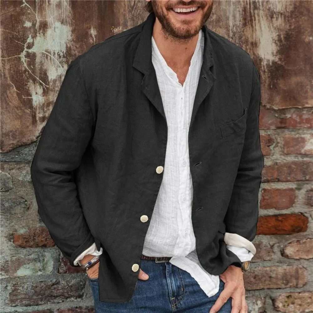 Man in zwarte linnen blazer met witte knoopsluiting, witte overhemd en blauwe jeans, leunend tegen een bakstenen muur. Casual herenkleding.