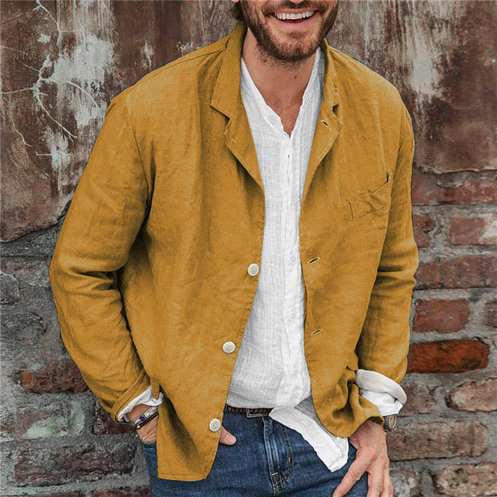 Man in gele linnen blazer, witte overhemd en blauwe jeans tegen een bakstenen muur. Stijlvolle casual herenkleding, modetrend 2023.