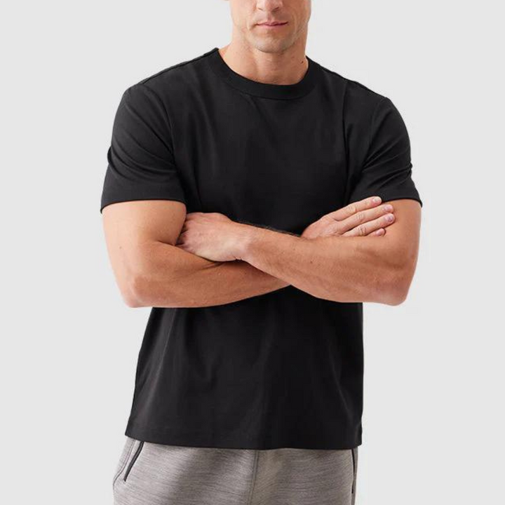 Man in zwarte T-shirt met gekruiste armen, grijze joggingbroek, neutrale achtergrond. Casual herenkleding, comfortabele stijl, modieuze outfit.