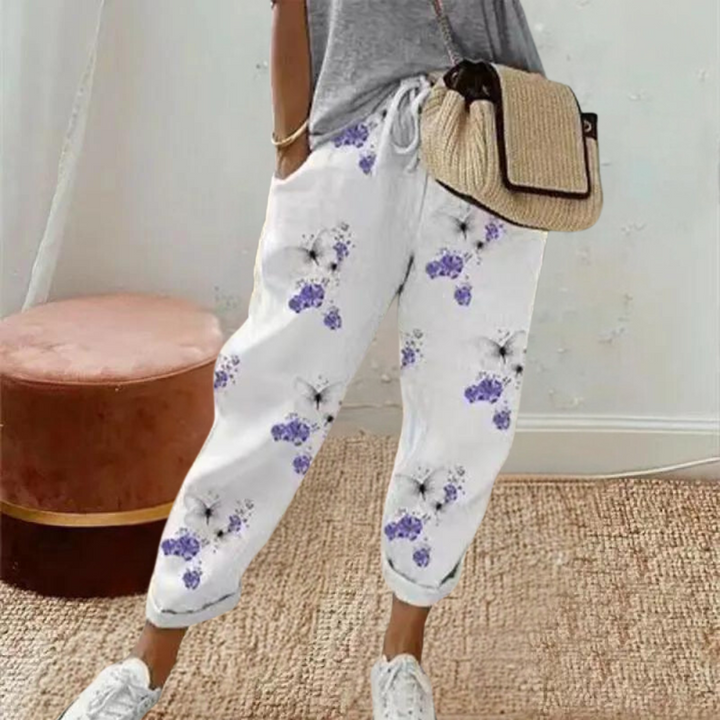 Vrouw draagt witte broek met paarse bloemenprint, gecombineerd met grijze top en rieten schoudertas. Trendy damesmode, casual outfit.