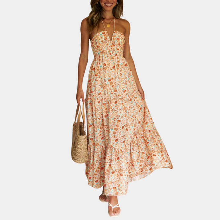 Vrouw in lange, oranje bloemenjurk met halternek en rieten tas. Zomerse damesmode, casual strandoutfit, trendy maxi-jurk.