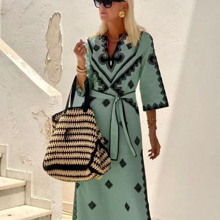 Vrouw in mintgroene jurk met zwarte patronen, draagt grote gevlochten tas en zonnebril, poseert bij witte muur. Zomermode, elegante stijl.