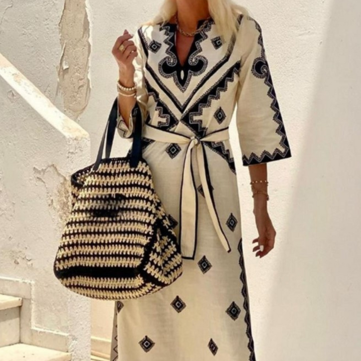 Vrouw in geborduurde beige jurk met zwarte patronen, draagt een grote, gevlochten tas. Stijlvolle zomermode, boho-chic, elegante outfit.