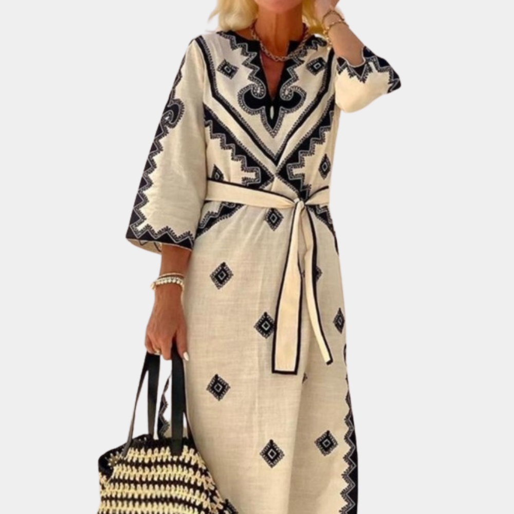 Vrouw in elegante, beige maxi-jurk met zwarte geometrische patronen en ceintuur, draagt een zwart-witte geweven tas. Perfect voor zomermode.