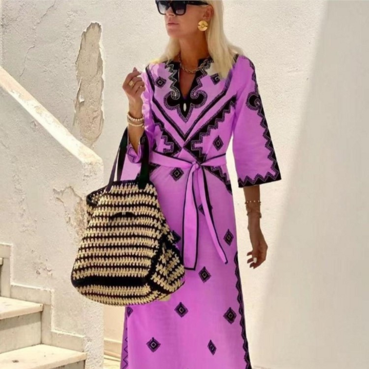 Vrouw in paarse boho-jurk met zwarte patronen, draagt grote gevlochten tas en zonnebril, poseert op trap. Zomermode, bohemian stijl.