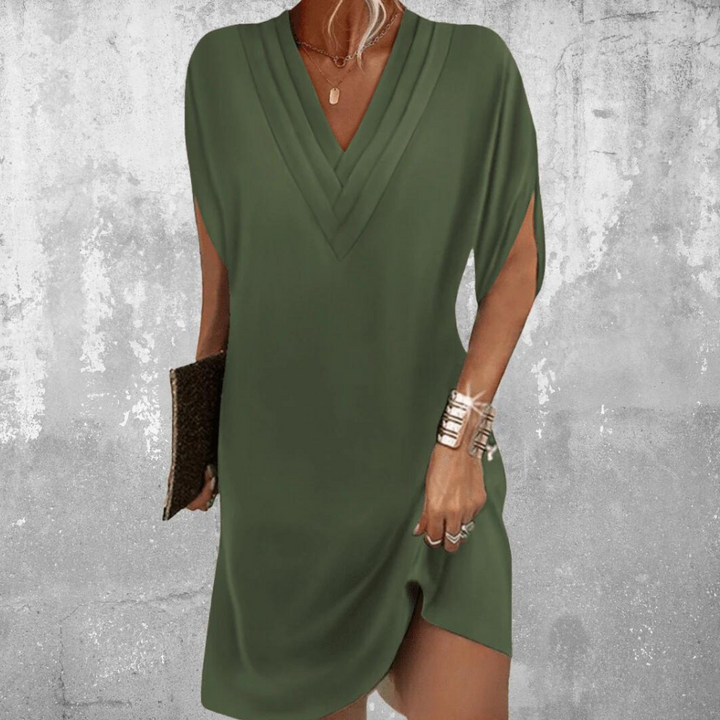 Vrouw in stijlvolle legergroene jurk met V-hals, asymmetrische zoom en zilveren armbanden, tegen een betonnen achtergrond. Perfect voor casual mode.