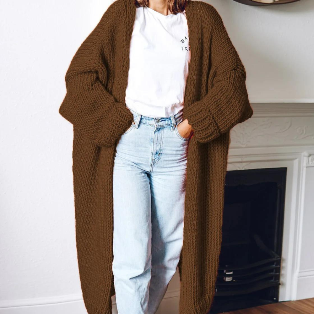 Vrouw draagt lange, bruine gebreide cardigan over witte T-shirt en blauwe jeans, staand voor open haard. Comfortabele herfstmode, casual stijl.