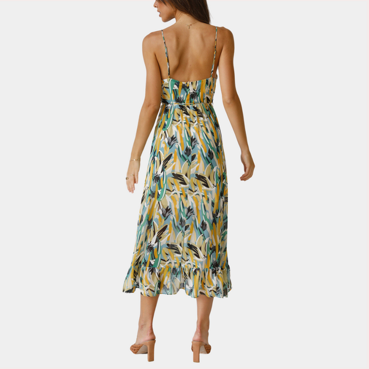 Vrouw in zomerse, mouwloze maxi-jurk met groene en gele bladerenprint, rugdecolleté, en ruches. Perfect voor casual en strandmode.