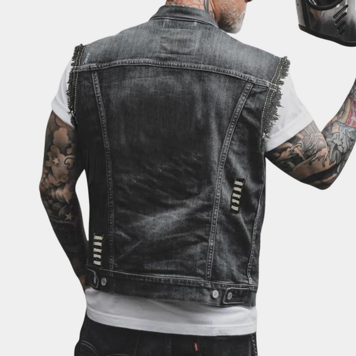 Man in zwart denim spijkerjack zonder mouwen, met tatoeages op armen, houdt motorhelm vast. Modieuze herenkleding, casual streetwear.