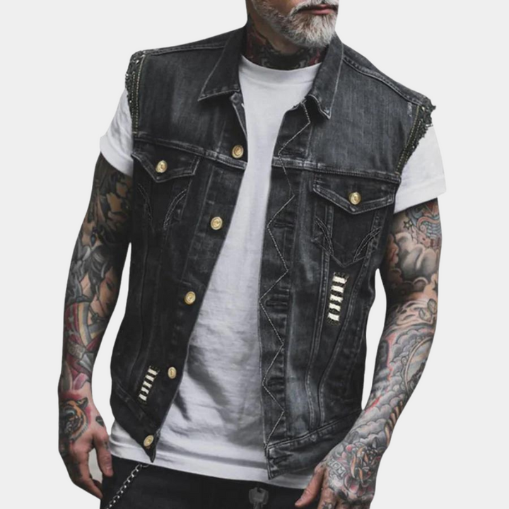 Man met tatoeages draagt een zwarte spijkerhesje met gouden knopen en witte T-shirt. Modieuze herenkleding, casual streetwear stijl.