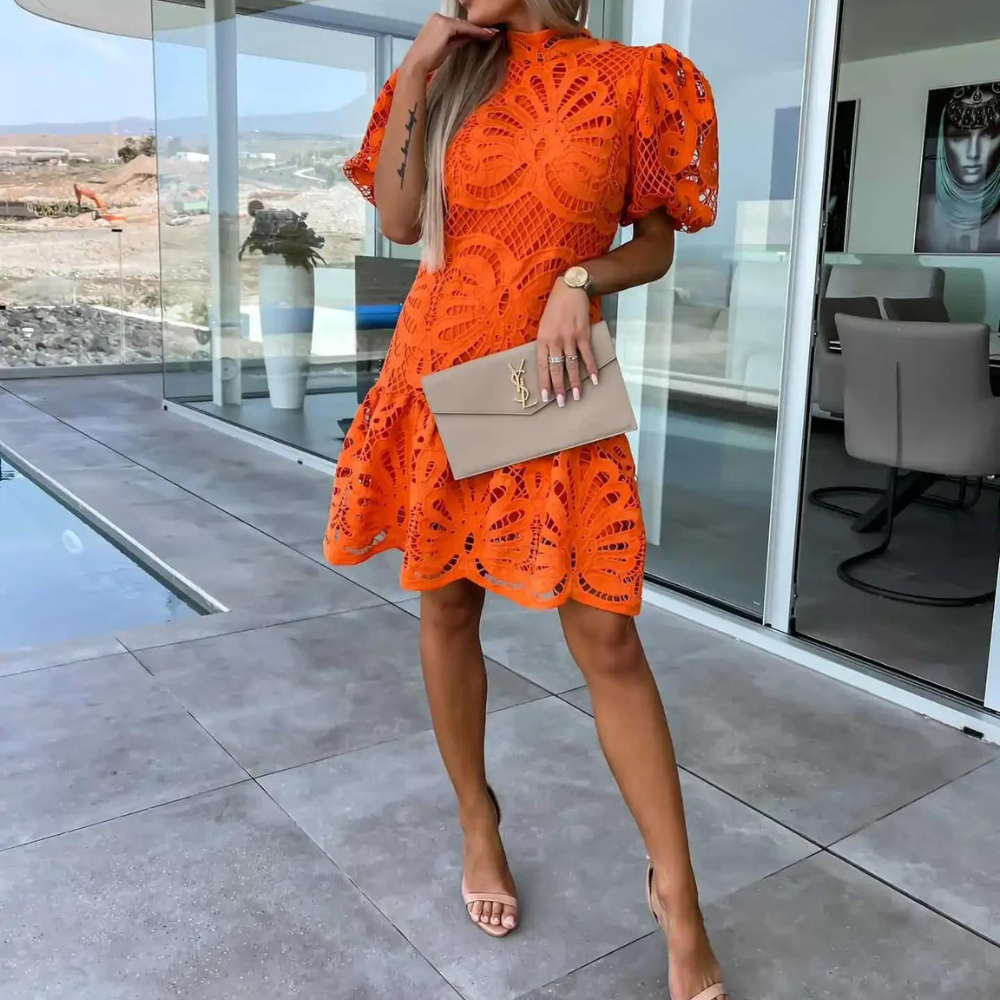 Oranje kanten jurk met korte mouwen, gedragen door vrouw met beige clutch en sandalen, staand op terras bij glazen deur. Zomerjurk, modetrend.