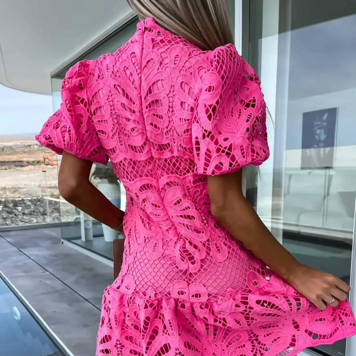 Vrouw in roze kanten jurk met pofmouwen, staand op balkon met glazen balustrade. Trendy zomerjurk, modieuze dameskleding, buitenomgeving.