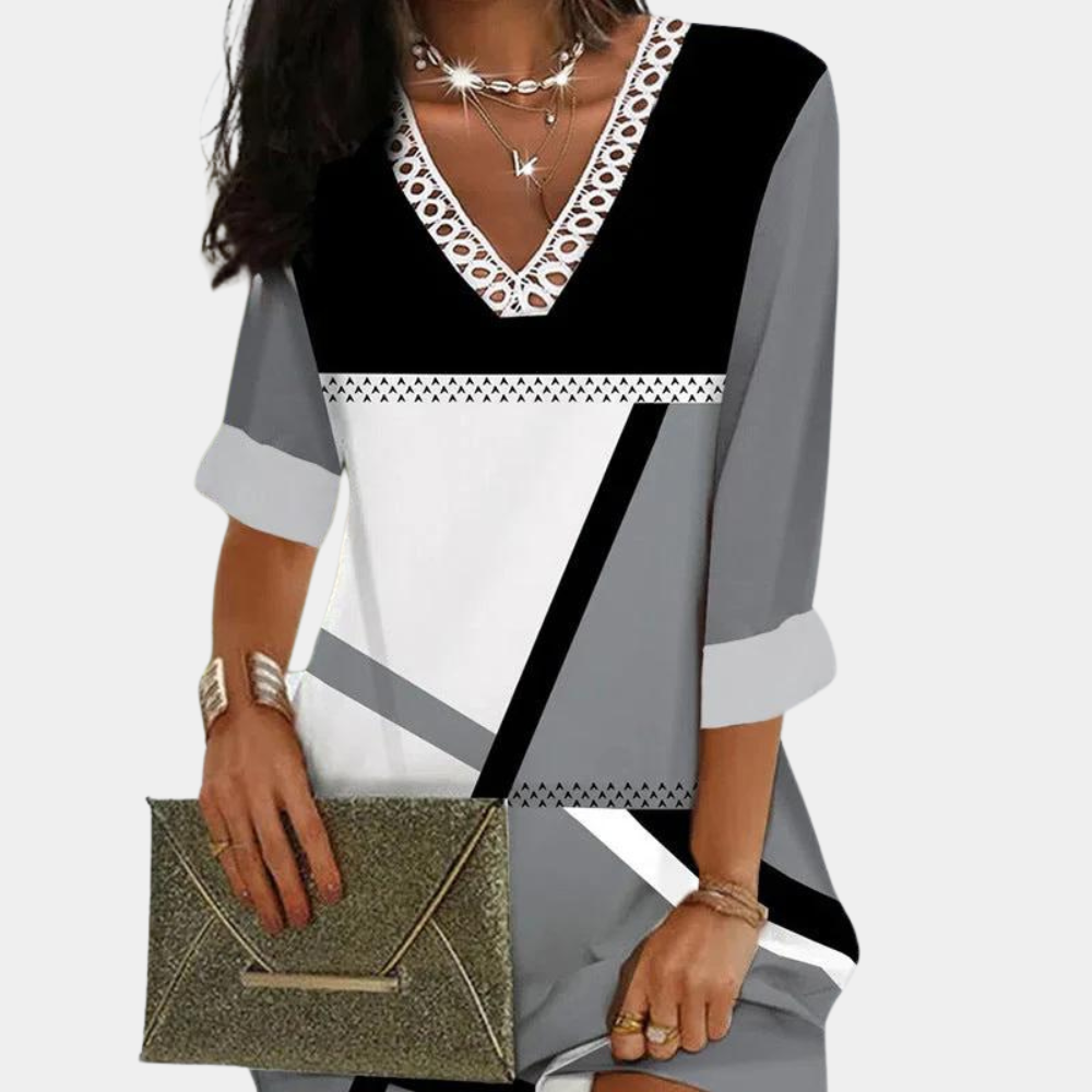 Vrouw in stijlvolle grijze en zwarte geometrische jurk met V-hals en kanten details, draagt een gouden clutch. Elegante mode, moderne dameskleding.