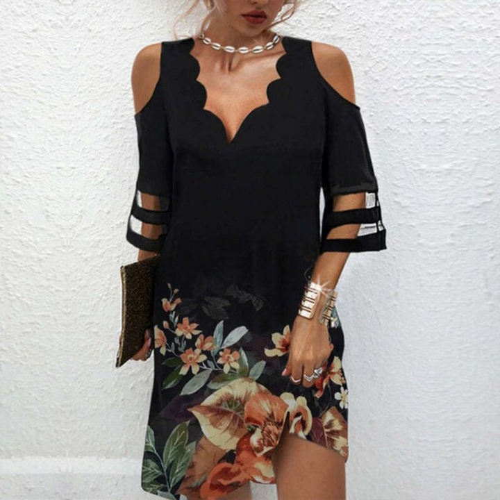 Vrouw in zwarte off-shoulder jurk met bloemenprint, elegante halslijn en driekwart mouwen, draagt een parelketting en gouden armband.