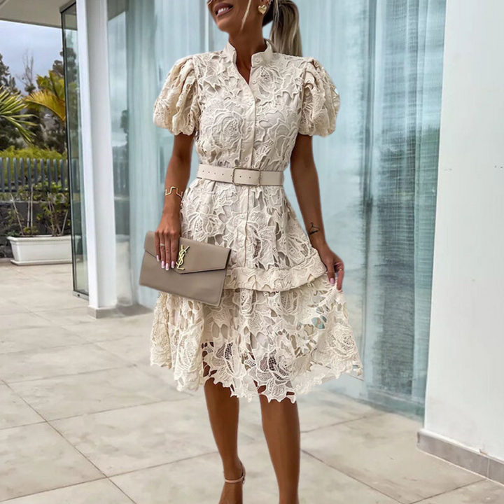 Vrouw in elegante kanten jurk met pofmouwen en riem, houdt beige clutch vast. Perfecte outfit voor zomerse evenementen. Luxe mode, dameskleding.
