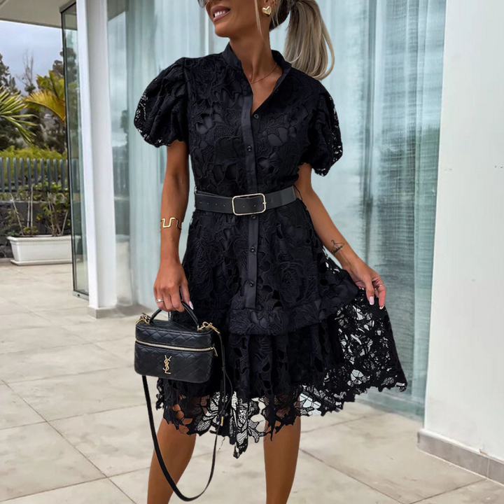 Elegante zwarte kanten jurk met korte pofmouwen en riem, gedragen door vrouw op terras. Perfect voor zomerfeestjes en modieuze evenementen.