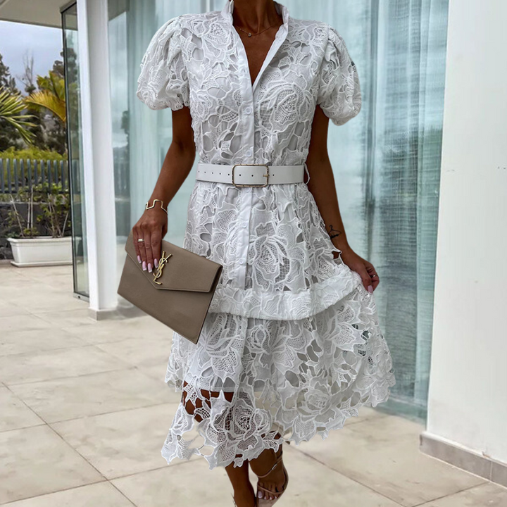 Vrouw in elegante witte kanten jurk met pofmouwen en riem, houdt een beige clutch. Perfect voor zomerse evenementen en modieuze gelegenheden.