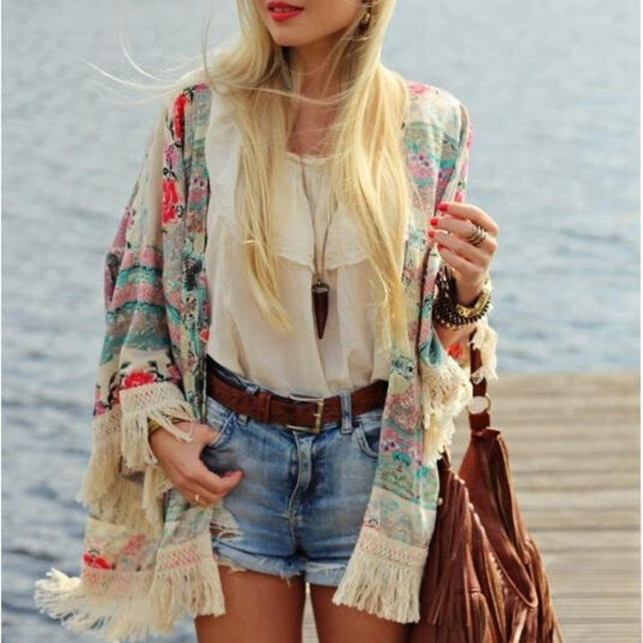 Vrouw in boho-stijl met bloemenkimono, witte blouse en denim shorts bij water. Trendy zomeroutfit, modeaccessoires, casual strandlook.