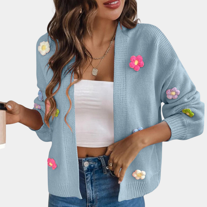 Vrouw draagt lichtblauw gebreid vest met kleurrijke bloemen, witte top en jeans. Trendy damesmode, casual stijl, herfstkleding.