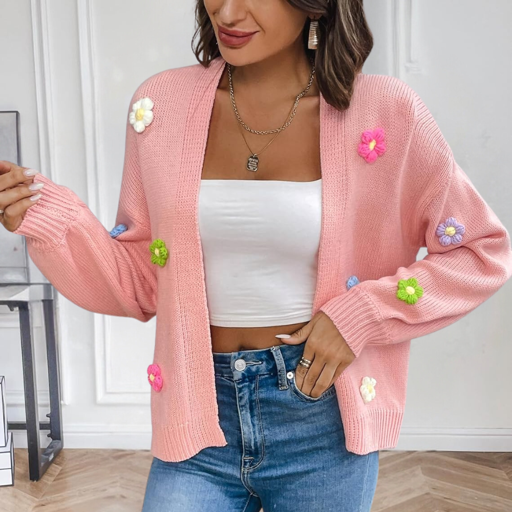 Vrouw draagt roze gebreid vest met kleurrijke bloemen, witte crop top en blauwe jeans. Trendy damesmode, casual outfit, herfstkleding.