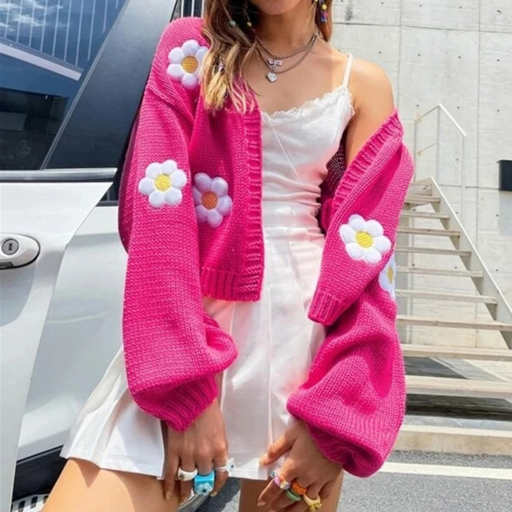 Vrouw in roze gebreid vest met bloemenpatroon, witte jurk, kleurrijke ringen, poseert buiten. Trendy damesmode, streetwear, zomeroutfit.