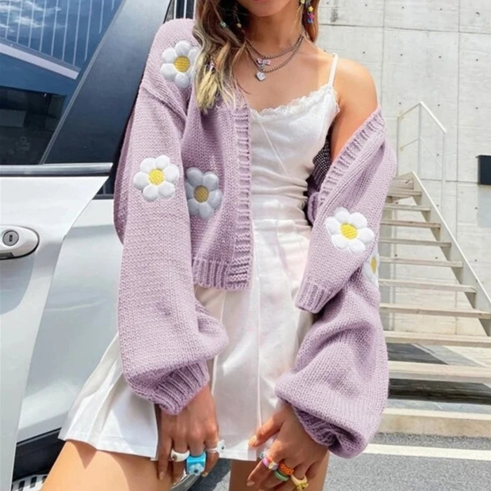 Vrouw in witte jurk met lila gebreid vest met madeliefjes, kleurrijke ringen, voor een auto. Trendy mode, zomerse outfit, straatstijl.